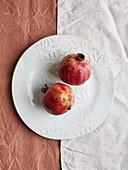 Zwei reife Granatäpfel liegen auf einem kunstvoll gestalteten weißen Teller vor einem kontrastierenden Stoffhintergrund. Die Komposition verbindet rustikalen Charme mit einem Hauch von natürlicher Farbe.