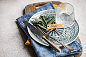 A sophisticated table setting with olive leaves on a ceramic plate with a shield, a glass and fine dining utensils.