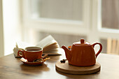 A cosy interior with an orange teapot on a wooden tray, a matching cup of tea and an open book on a table. The sunlight shines through a nearby window and intensifies the warmth.