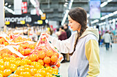 In a busy supermarket, a woman selects oranges from the fresh produce section and carefully examines a bag of fruit.