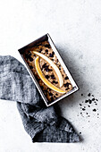A loaf of banana bread, topped with banana slices and chocolate chips, lying on a grey linen cloth against a light background.