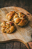 Three freshly baked, golden-brown cardamom buns on a wooden cutting board. The background is a dark brown wooden table, which gives the picture a warm and inviting look and evokes a feeling of comfort and enjoyment.