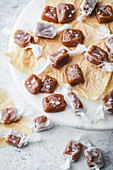 A close-up of homemade caramel sweets on a marble surface. The candies are wrapped in white parchment paper and sprinkled with sea salt. They are arranged on a piece of brown parchment paper standing on a white marble cake stand. More caramels can be seen in the blurred background, hinting at a delicious treat and highlighting the simplicity and beauty of homemade sweets.