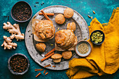 A colourful arrangement of autumnal chai spices and fresh, golden brown muffins on an antique pewter plate. Various spices in bowls, as well as fresh ginger and cinnamon. A bright saffron yellow linen cloth provides a colour accent. The background consists of a custom-made blue-grey textured surface that provides a colourful, sophisticated backdrop for the warm, autumnal tones of the spices and baked treats.