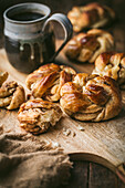 Ein rustikales Holzbrett ist mit frisch gebackenen Kardamombrötchen beladen, deren goldbraune Oberfläche mit einer süßen Glasur glänzt. Die Brötchen sind verführerisch aufgereiht, einige sind aufgeklappt und geben das weiche, fluffige Innere preis. Neben den Brötchen steht ein rustikaler Keramikbecher, gefüllt mit dunklem Kaffee. Die warmen, einladenden Töne des Bildes erwecken ein Gefühl von Gemütlichkeit und Genuss, wobei der Schwerpunkt auf der Köstlichkeit der Backwaren und dem Vergnügen eines einfachen Morgenrituals liegt.
