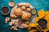 A vibrant, flat layer of autumnal chai spices and fresh, golden brown muffins on an antique pewter plate. Various spices in bowls, as well as fresh ginger and cinnamon. A bright saffron yellow linen cloth provides a colour accent. The background consists of a custom-made blue-grey textured surface that provides a colourful, sophisticated backdrop for the warm, autumnal tones of the spices and baked treats.