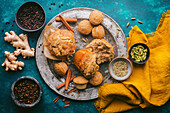 A rustic, teal-coloured backdrop with a variety of chai spices and freshly baked muffins, some of which are whole and others broken apart to reveal their fluffy insides. Around the plate are cinnamon sticks, fresh ginger and small bowls of various spices, including black peppercorns, cloves, fennel seeds and cardamom pods, adding a touch of natural colour and texture. A saffron-coloured linen napkin adds a warm accent and complements the overall rustic aesthetic. The picture captures a simple but