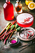 Beautiful pink-coloured cocktail with a frothy rim in a coupe glass with a simple rhubarb curl garnish, fresh rhubarb stalks and syrup in the background