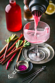 Action shot of a pink rhubarb cocktail poured from the shaker into a beautiful coupe glass with fresh rhubarb and red syrup in the background