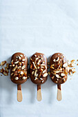 Three almond ice cream sticks covered in milk chocolate, sprinkled with roasted almonds on a white marble background