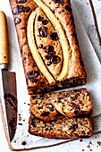 A sliced banana chocolate chip bread with a knife next to it, which has a moist texture and is topped with fresh banana slices.