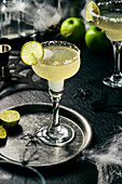 Lime margarita with salt rim on a black surface, Halloween setting, lime slices on a black tray