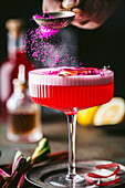 An action shot of bright pink berry powder sprinkled on a sophisticated alcoholic drink with a textured, stemmed coupe cocktail glass with colourful red liqueur and a frothy top