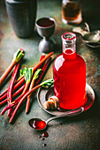 Beautiful glass bottle with crimson rhubarb syrup and syrup in a spoon, with fresh rhubarb stalks and cocktail jigger in the background