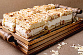 Homemade Norwegian sweet cake Verdens Beste Kake with pudding, meringue and almonds, cut into squares, served on a wooden tray. Beige-coloured table, close-up.