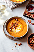 A bowl of creamy carrot soup, garnished with red pepper strips, crispy chickpeas and a cream swirl, served on a rustic plate.