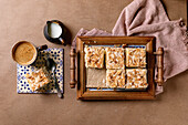 Homemade Norwegian sweet cake Verdens Beste Kake with pudding, meringue and almonds, cut into squares, served on a wooden tray. Beige background, pink fabric, coffee cup, spoon, milk jug. View from above
