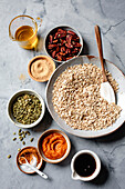 A selection of ingredients for a muesli recipe, including rolled oats, pecans, pumpkin puree, pumpkin seeds, brown sugar, spices, maple syrup and oil on a marble worktop.