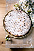 Homemade Pastiera Napolitano cake dusted with icing sugar on a rustic wooden table with white spring flowers. Traditional pastry with flower-shaped decorations.