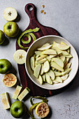 A selection of green apples, some sliced and placed in a bowl, together with apple peels and a halved lemon, all arranged on a wooden chopping board.