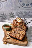 Sliced homemade banana bread with walnuts, drizzled with caramel and topped with fresh banana. Placed on a wooden board with a bowl of sauce on a white marble base. View from above