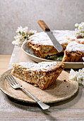 Homemade Pastiera Napolitano cake in slices with icing sugar on a ceramic plate. Serving knife, fork, white flowers. Rustic wooden background. Delicious dessert.