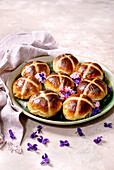 Traditional hot cross buns on a ceramic plate decorated with edible flowers. Homemade Easter buns on a light-coloured background with linen napkin and purple petals. Close-up