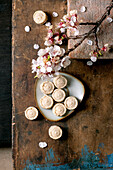 Homemade sesame cream sweets on a white plate with a branch of cherry blossom in bloom. Rustic wooden background with spring flowers and pastries. Top view of stylish composition.