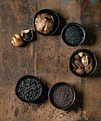 Set of five black spices: black fermented garlic, black Himalayan salt, black mustard seeds, black cardamom and fermented campota pepper in black ceramic bowls. Dark wooden background. Close-up