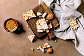 Traditional Spanish dessert: crispy turron with almonds, broken into pieces on a wooden board. Linen napkin, espresso coffee in a black cup. Beige-coloured textured background, view from above.