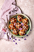 Hot cross buns on a ceramic plate garnished with pink and purple flowers. Traditional Easter pastries on a light coloured textured background with pink linen napkin. Festive spring holiday baking view from above