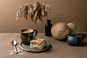 Homemade Norwegian sweet cake Verdens Beste Kake with vanilla pudding, meringue and almonds, a square piece on a plate on a beige table with coffee cup, spoon, milk jug, reed in bottle, beige wall.