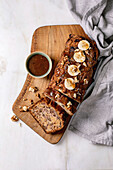 Sliced homemade banana bread with walnuts, drizzled with caramel and topped with fresh banana. Placed on a wooden board with a bowl of sauce on a white marble base. View from above