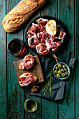 Sliced Spanish ham Jamon Iberico paleta on a black plate. Bowl of green olives, fresh and toasted white bread, olive oil, glass of red wine, tapas. Old wooden background, dark turquoise colour. Place to copy