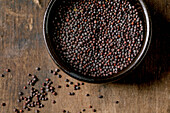 Black mustard seeds for cooking Dijon mustard in a black ceramic bowl on a dark wooden table. Healthy food, savoury cooking ingredients, traditional cuisine. Top view, close-up