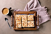 Homemade Norwegian sweet cake Verdens Beste Kake with pudding, meringue and almonds, cut into squares, served on a wooden tray. Beige background, pink fabric, coffee cup, spoon, milk jug. View from above