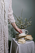 Woman in white linen dress arranges ceramic jug with spring flowers. Placemat with slices of sandwich and coffee cup against a grey background. Minimalist breakfast styling.