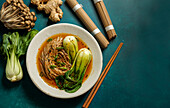 Bowl of soba noodles in a spicy broth with baby bok choy, surrounded by fresh mushrooms and ginger
