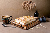 Homemade Norwegian cake Verdens Beste Kake with pudding, meringue and almonds, cut into squares, served on a wooden tray. Beige-coloured table with coffee cup, spoon, milk jug, dry reed in a bottle, beige-coloured wall.