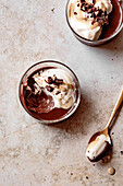 Two glass bowls of chocolate pudding with whipped cream and chocolate shavings, next to a spoon on a textured surface.