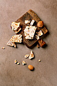 Traditional Spanish dessert, classic crunchy turron with almonds, broken into pieces on a wooden serving board. Beige-coloured, textured background. Top view, distance
