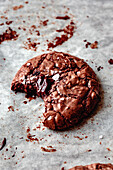 Close-up of a chocolate biscuit from which a piece has been bitten out, with sticky chocolate inside and sprinkled sea salt on top, lying on parchment paper.
