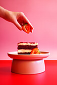 Female hand placing a strawberry on a slice of double layer red velvet cake. Pop art food colour blocking aesthetic.
