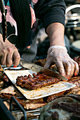 Straßenküchenfest in Europa. Traditionelle europäische gegrillte Schweinerippchen in Soße. Verkäuferin mit Handschuhen bereitet eine Portion Rippchen an einem Essensstand zu.