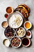 An assortment of ingredients for a recipe, including ripe bananas, various nuts, flour and spices, arranged on a textured surface.