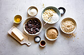 A selection of baking ingredients such as chocolate chips, butter, sugar, egg, flour, oat flakes and vanilla extract are arranged on a light-coloured surface.