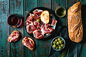 Sliced Spanish ham Jamon Iberico paleta on a black plate. Bowl of green olives, fresh and toasted white bread, olive oil, glass of red wine, tapas. Old wooden background, dark turquoise colour. Place to copy