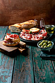 Sliced Spanish ham Jamon Iberico paleta on a black plate. Bowl of green olives, fresh and toasted white bread, olive oil, glass of red wine, tapas. Old wooden background, dark turquoise colour. Place to copy