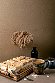 Homemade Norwegian cake Verdens Beste Kake with pudding, meringue and almonds, cut into squares, served on a wooden tray. Beige-coloured table with coffee cup, spoon, milk jug, dry reed in a bottle, beige-coloured wall.