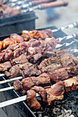 Nahaufnahme von marinierten Fleischstücken, die auf Metallspießen über einem heißen Holzkohlegrill gegart werden. Saftige Rindfleischspieße mit rauchigem Geschmack, perfekt für eine Grillparty oder zum Kochen im Freien.
